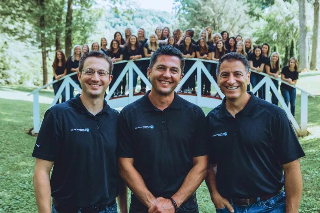 Three orthodontists in black polo shirts smiling in front of a group of staff on a white railing, surrounded by greenery, representing Holmes & Palmer Orthodontics in Charleston, WV.