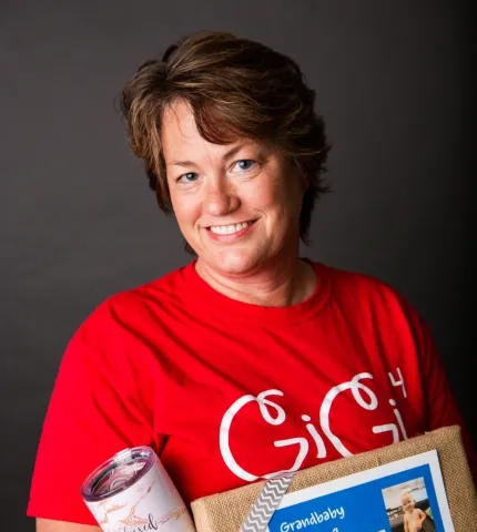 Woman smiling in a red "GiGi" t-shirt holding a photo of her grandchild and a tumbler, representing family and personal connection in the Holmes & Palmer Orthodontics team.