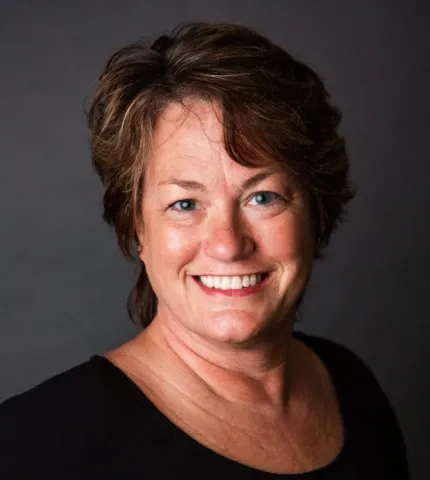 Shelli Diedrich, smiling Scheduling Coordinator Director at Holmes & Palmer Orthodontics, with short brown hair and wearing a black top, against a dark background.